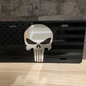 May include: Black and white metal license plate featuring a skull with a white American flag background.