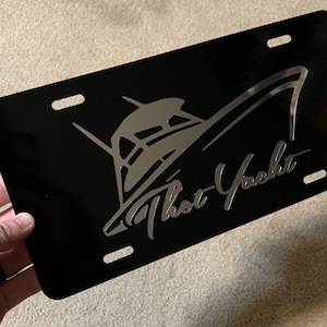 May include: Black license plate with a silver boat graphic and the text "That Yacht". The plate has four mounting holes and a glossy finish. The design features a stylized boat.