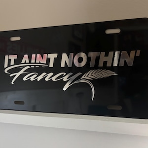 May include: Black metal license plate with silver lettering that reads "It ain't nothin' Fancy" with a wheat stalk graphic.