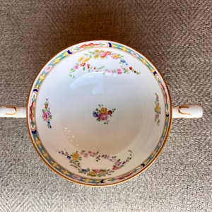 May include: A white porcelain soup bowl with two handles, decorated with a floral pattern in pink, yellow, and blue. The rim has a gold and teal border. The bowl is designed for serving soups and stews.