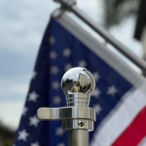 May include: Close-up of a shiny, chrome-finished flagpole hardware. The hardware includes a spherical finial, a cleat, and a pole. The American flag with red and white stripes and blue field with white stars is in the background.