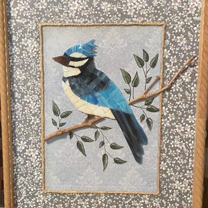 Può includere: Un'opera d'arte a mosaico incorniciata raffigurante una ghiandaia azzurra realizzata con pezzi di vetro blu, neri e bianchi. L'uccello è appollaiato su un rametto naturale con foglie verdi su uno sfondo azzurro, il tutto in una cornice di legno chiaro.
