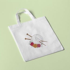 May include: White tote bag with a black and white illustration of a ball of yarn with knitting needles and a floral wreath. The flowers are red, pink, and orange.