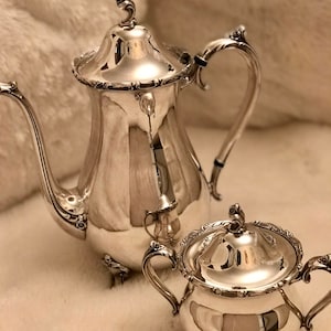 May include: A silver tea set with a teapot and a sugar bowl. Both pieces have ornate designs and are polished to a shine.