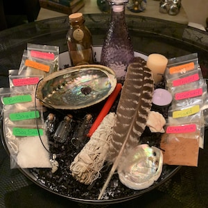 May include: A black bowl filled with various ingredients for a spell or ritual. The ingredients include sea salt, sage, basil, Himalayan pink salt, bay leaves, cinnamon, sand, rosemary, thyme, and a feather. There are also two small glass bottles, a purple glass bottle with a stopper, a red candle, and a white sage smudge stick.