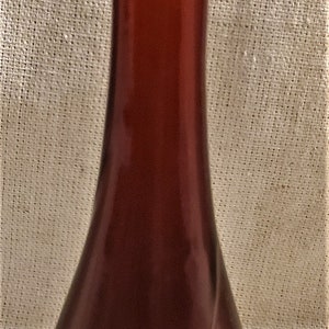 Könnte beinhalten: Eine dunkelrote Glasflasche mit einem runden, dunkelbraunen Stopfen. Die Flasche hat einen breiten Boden und einen schmalen Hals.