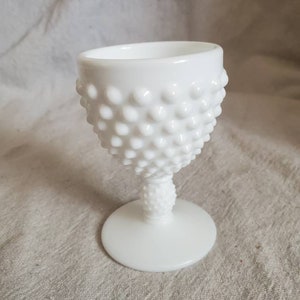 Könnte beinhalten: Ein weißes Hobnail-Glasbecher mit rundem Fuß und einem strukturierten, abgerundeten Körper. Der Becher hat einen kurzen Stiel und einen glatten Rand. Das Hobnail-Design bedeckt den Körper des Glases und erzeugt eine holprige Textur. Das Glas ist undurchsichtig und scheint aus Milchglas zu sein.