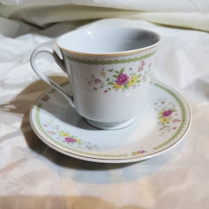 Puede incluir: Juego de taza y platillo blancos con detalles florales y dorados. La taza tiene un asa curvada y está decorada con flores rosas, amarillas y moradas. El platillo tiene un diseño floral a juego y un borde dorado. Un juego de té elegante.