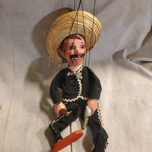 May include: A vintage string puppet of a man wearing a straw sombrero, black pants with white trim, and red shoes. He has a mustache and is holding a gun in one hand.