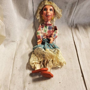 May include: A vintage hand puppet with a red and white checkered shirt, a blue and white patterned skirt, and a white lace hat. The puppet has a painted face with red lips and is wearing red shoes.