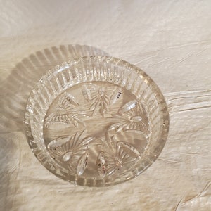 Può includere: Un sottobicchiere di vetro trasparente con un motivo decorativo a forma di stella. Il sottobicchiere ha un bordo rialzato e una finitura liscia e lucida.