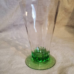 May include: A clear glass drinking glass with a green base and a textured design. The glass has a wide mouth and a narrow base.