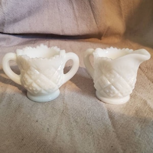 Könnte beinhalten: Ein weißes Milchglas-Sahnegießkanne und Zuckerdose-Set mit einem Diamantenmuster. Die Kanne hat einen Griff und einen Ausguss. Die Zuckerdose hat zwei Griffe.