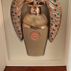 May include: A beige perfume bottle with gold detailing and a decorative peacock design. The peacock feathers are embellished with colourful jewels. The bottle is presented in a white box. The bottle has text on it.