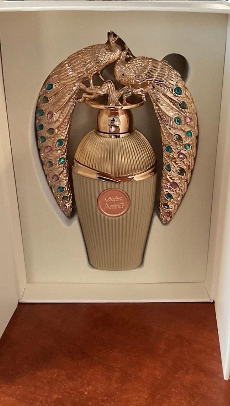 May include: A beige perfume bottle with gold detailing and a decorative peacock design. The peacock feathers are embellished with colourful jewels. The bottle is presented in a white box. The bottle has text on it.
