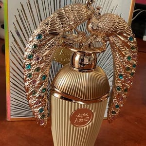 May include: A gold-coloured perfume bottle with a decorative peacock design. The bottle has a ribbed, cream-coloured base and a gold cap. The peacock design features colourful gemstones. The packaging has the text "Areef" on the front.