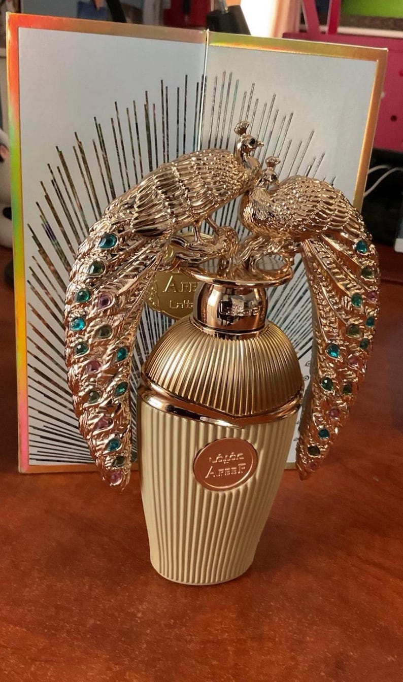 May include: A gold-coloured perfume bottle with a decorative peacock design. The bottle has a ribbed, cream-coloured base and a gold cap. The peacock design features colourful gemstones. The packaging has the text "Areef" on the front.