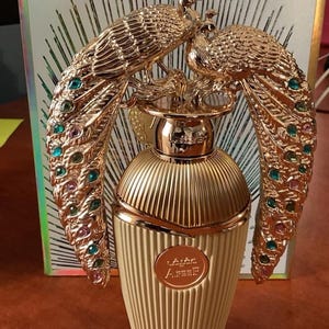 May include: A gold perfume bottle with a peacock design. The bottle has a ribbed texture and a gold cap. The peacock design features colourful jewels. The perfume bottle is set against a background with a geometric pattern.