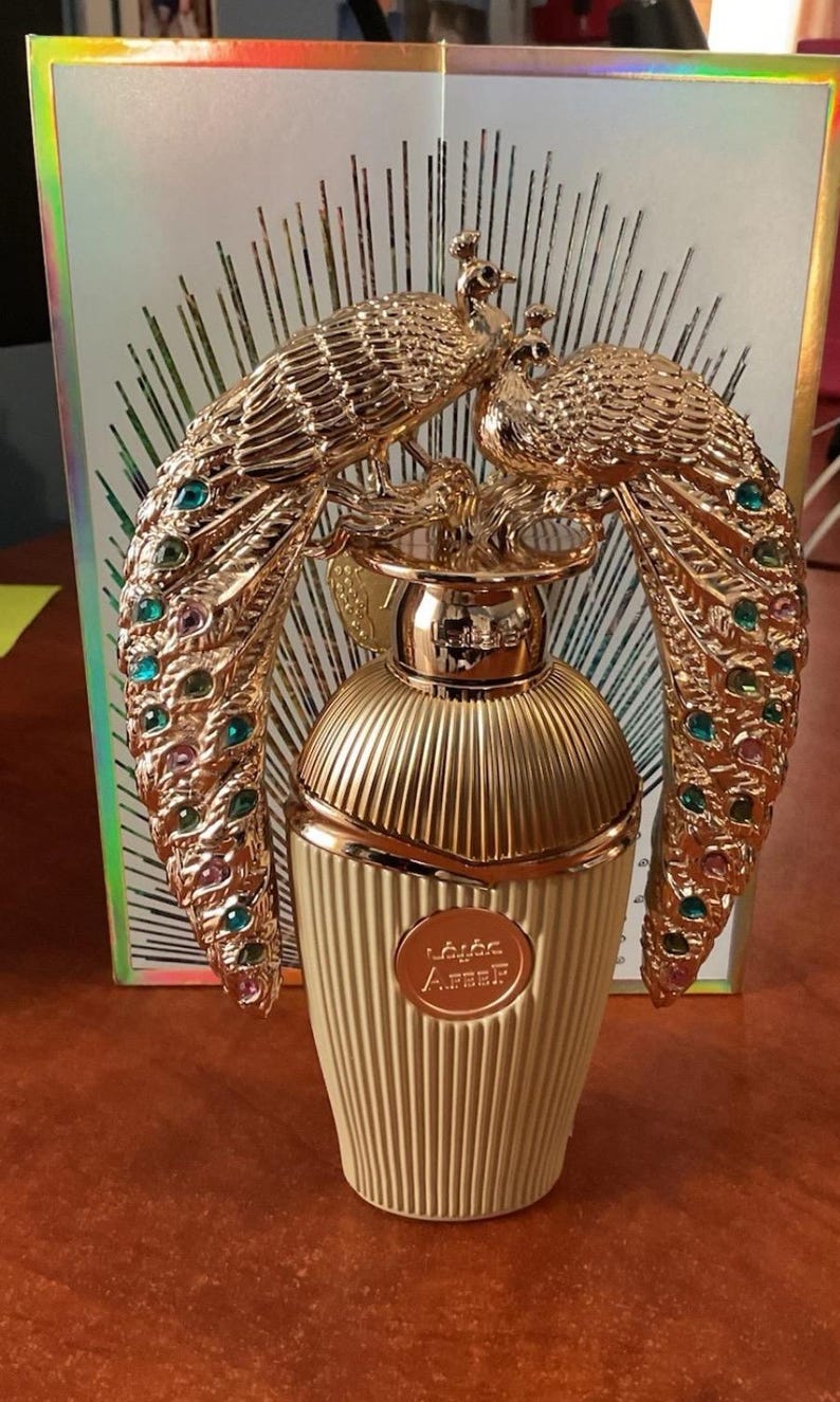 May include: A gold perfume bottle with a peacock design. The bottle has a ribbed texture and a gold cap. The peacock design features colourful jewels. The perfume bottle is set against a background with a geometric pattern.