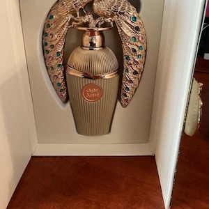 May include: A perfume bottle with a gold peacock design. The bottle is light tan with vertical ridges. The peacock design is gold with colourful accents. The perfume bottle is presented in a white box.