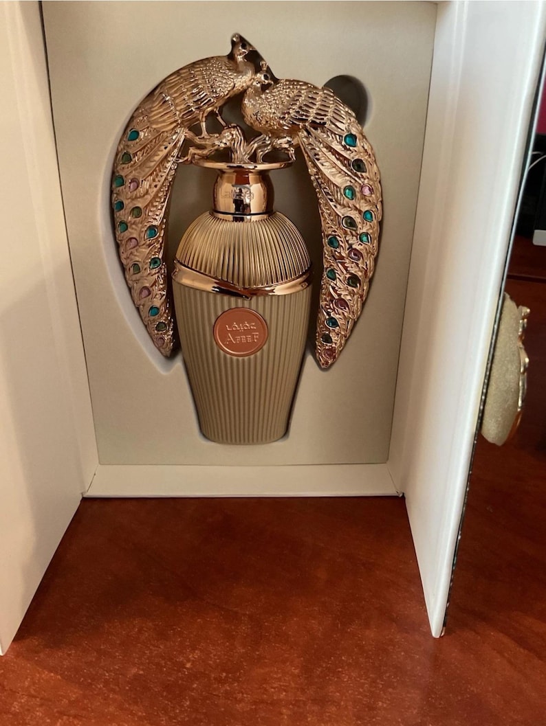 May include: A perfume bottle with a gold peacock design. The bottle is light tan with vertical ridges. The peacock design is gold with colourful accents. The perfume bottle is presented in a white box.