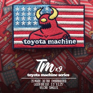 May include: A rectangular embroidered patch featuring a red horned character over a blue and white star-spangled banner design with the text toyota machine. The patch measures 3.5 inches by 2.25 inches and includes a velcro backing for attachment.