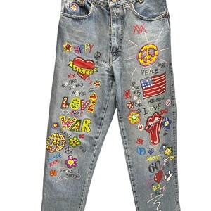 Könnte beinhalten: Hellblaue Jeans mit farbenfrohen, handgezeichneten Grafiken. Die Jeans zeigen die Wörter "Love", "Peace", "War" und "Sixty" sowie ein Friedenszeichen, ein Herz und eine amerikanische Flagge. Verziert mit Blumen und anderen Symbolen.