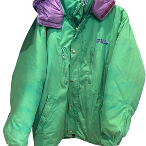 Può includere: Giacca invernale verde con cappuccio e colletto viola. La giacca ha una chiusura a cerniera, più tasche e il logo Fila ricamato sul petto. La giacca è appesa a un gancio di metallo.