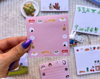 May include: A square pink sticky note featuring a border of small food illustrations including sushi, boba tea, fried shrimp, and eggs. The text @NESSANDESIGN is printed at the bottom center. The note is held against a background of other stationery.
