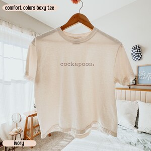May include: An ivory short-sleeve boxy t-shirt with a crew neckline. The center of the chest features the text 'cockapoos.' printed in a dark gray, lowercase sans-serif font. The garment is displayed on a wooden hanger against a light background.
