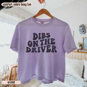 K&ouml;nnte beinhalten: Ein helllila Comfort Colors Boxy T-Shirt mit dem Text "Dibs on the Driver" in Dunkelgrau. Das Shirt h&auml;ngt an einem Holzkleiderb&uuml;gel. Das Wort "orchid" ist auf einem Etikett am unteren Rand des Bildes aufgedruckt.