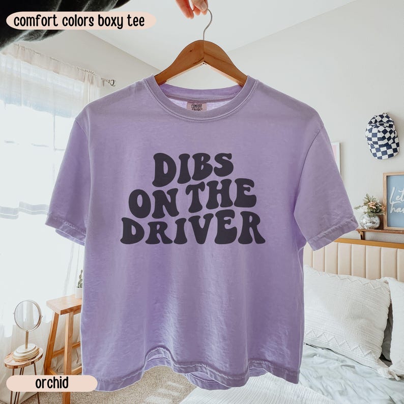 K&ouml;nnte beinhalten: Ein helllila Comfort Colors Boxy T-Shirt mit dem Text "Dibs on the Driver" in Dunkelgrau. Das Shirt h&auml;ngt an einem Holzkleiderb&uuml;gel. Das Wort "orchid" ist auf einem Etikett am unteren Rand des Bildes aufgedruckt.