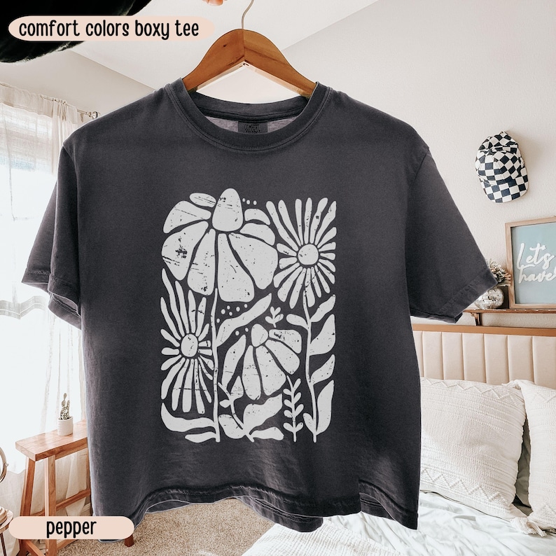 以下が含まれることがあります： 白の花柄がプリントされた、濃いグレーのComfort Colors boxy Tシャツ。クルーネックと半袖です。画像の下部には「pepper」の文字があります。