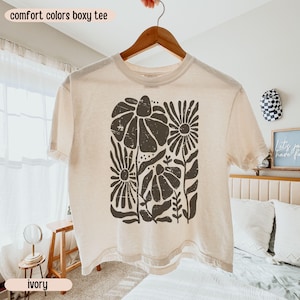 以下が含まれることがあります： アイボリー色のボクシーTシャツ。濃い茶色の花柄のデザイン。木製のハンガーにTシャツが掛かっています。「comfort colors boxy tee」の文字が上に、「ivory」の文字が下にあります。