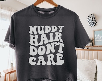May include: A dark gray boxy tee shirt with the phrase "MUDDY HAIR DON'T CARE" in white, retro-style lettering. The shirt is hanging on a wooden hanger. The word "pepper" is written on a label at the bottom.