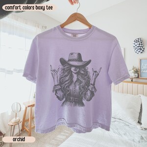 Peut inclure: Un t-shirt boxy violet clair avec une illustration graphique gris fonc&eacute; repr&eacute;sentant une personne aux longs cheveux ondul&eacute;s portant un chapeau de cowboy et faisant des gestes de la main rock and roll. Le t-shirt a un col rond et des manches courtes, avec le texte comfort colors boxy tee et orchid imprim&eacute; sur l'image.