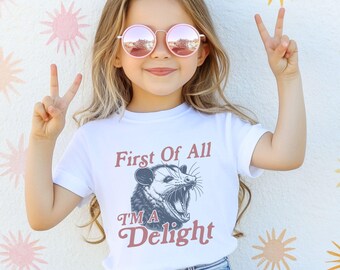 May include: White t-shirt with the text "First Of All I'm A Delight" and a graphic of an opossum. The person in the image is wearing pink sunglasses and making a peace sign with their fingers.