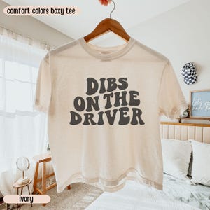 K&ouml;nnte beinhalten: Elfenbeinfarbenes, kastenf&ouml;rmiges T-Shirt mit dem Text "DIBS ON THE DRIVER" in Dunkelgrau, pr&auml;sentiert auf einem Holzb&uuml;gel. Die W&ouml;rter "comfort colors boxy tee" und "ivory" sind ebenfalls sichtbar.