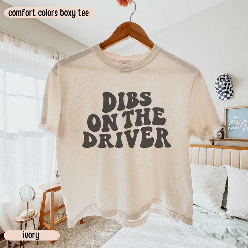 K&ouml;nnte beinhalten: Elfenbeinfarbenes, kastenf&ouml;rmiges T-Shirt mit dem Text "DIBS ON THE DRIVER" in Dunkelgrau, pr&auml;sentiert auf einem Holzb&uuml;gel. Die W&ouml;rter "comfort colors boxy tee" und "ivory" sind ebenfalls sichtbar.