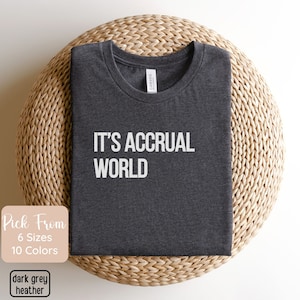 May include: Dark grey heather t-shirt with the text "IT'S ACCRUAL WORLD" in white. The shirt is folded and displayed on a woven, circular surface. Additional text indicates the availability of multiple sizes and colors.
