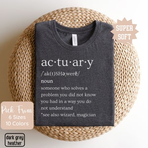 May include: Dark grey heather t-shirt with the definition of "actuary" printed in white. The definition includes the pronunciation, the word "noun", and a description of what an actuary does. The shirt is folded on a woven surface.