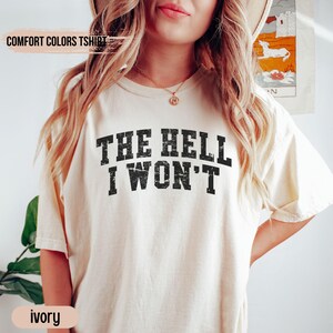 May include: A white t-shirt with the text "THE HELL I WON'T" printed in black. The shirt is a relaxed fit and has a distressed look.