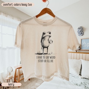 May include: Ivory-colored Comfort Colors boxy tee with a black and white illustration of a frog holding a mug. The text "I HAVE TO SAY WEIRD STUFF OR I'LL DIE" is printed below the frog.