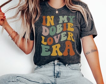 May include: Dark gray t-shirt with the text "IN MY DOBIE LOVER ERA" in retro-style lettering. The text is in shades of orange, brown, green, and pink. The shirt is paired with light blue jeans.