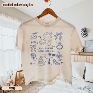 May include: Ivory-colored boxy tee shirt with a blue graphic design. The design includes cherries, doves, a seashell, a swimsuit, a ring, bows, flowers, a cake, a starfish, sunglasses, lipstick, a high heel, and the text "OLIVIA'S BACH CHARLESTON, SC".