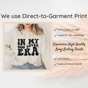 May include: A cream-colored sweatshirt with the text "IN MY DOG LOVER ERA" printed in black. The text is in a retro font style. The sweatshirt is being modeled by a person.