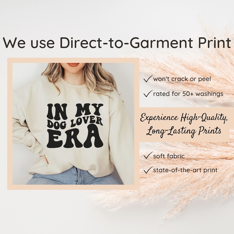 May include: A cream-colored sweatshirt with the text "IN MY DOG LOVER ERA" printed in black. The text is in a retro font style. The sweatshirt is being modeled by a person.