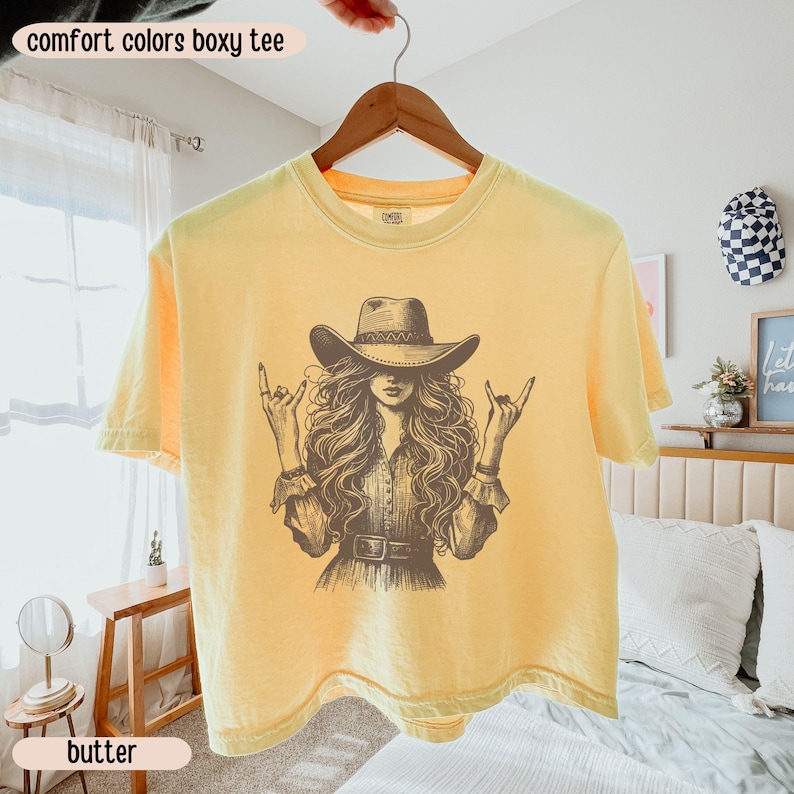 Peut inclure: Un t-shirt jaune p&acirc;le &agrave; coupe carr&eacute;e avec une impression graphique marron fonc&eacute; repr&eacute;sentant une personne aux longs cheveux ondul&eacute;s portant un chapeau de cowboy et faisant un signe de la main rock and roll. Le texte comfort colors boxy tee est pr&eacute;sent.