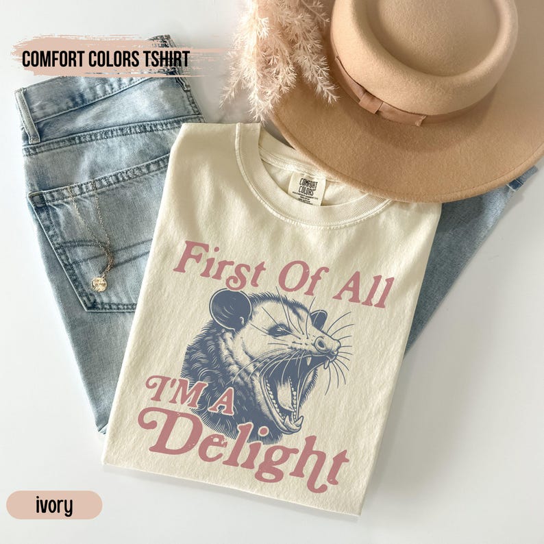 May include: Ivory t-shirt with a graphic of a possum with the text "First of all I'm a Delight" in pink and blue lettering.