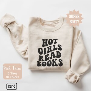 May include: A beige sweatshirt with the text "HOT GIRLS READ BOOKS" in black, retro-style lettering. The sweatshirt is folded and laid flat on a white surface.